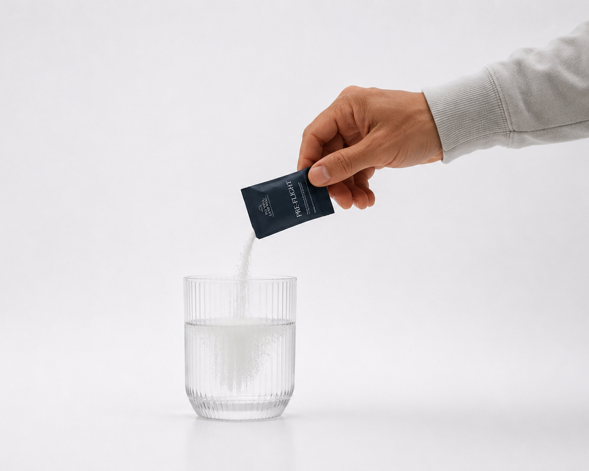 A hand in a light gray long-sleeve shirt pours white powder from a small dark blue packet into a clear ribbed glass of water, set against a seamless white background.
