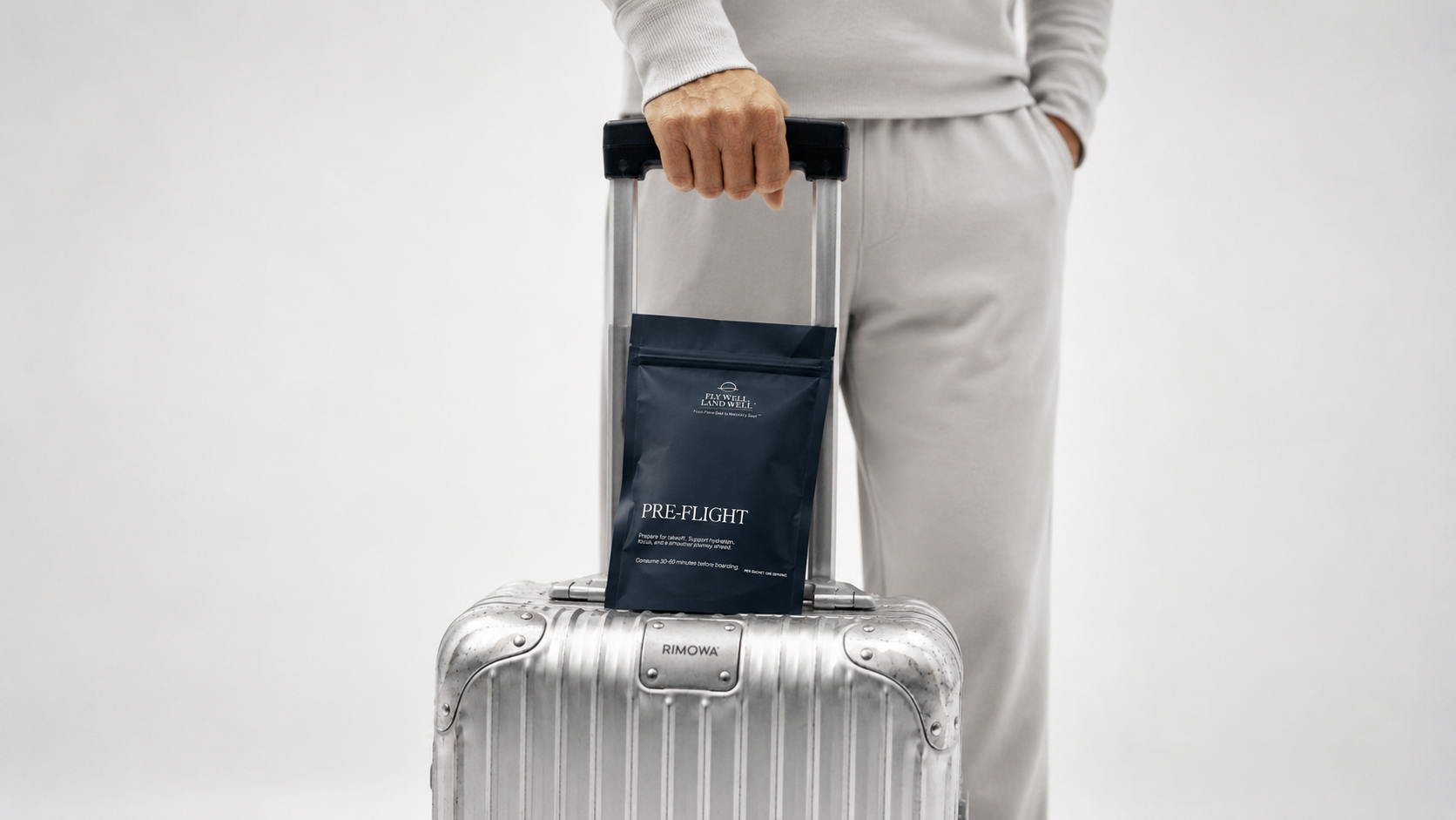 A person in light gray loungewear holds the handle of a silver rolling suitcase while standing next to a dark navy blue pouch labeled PRE-FLIGHT and Rimowa