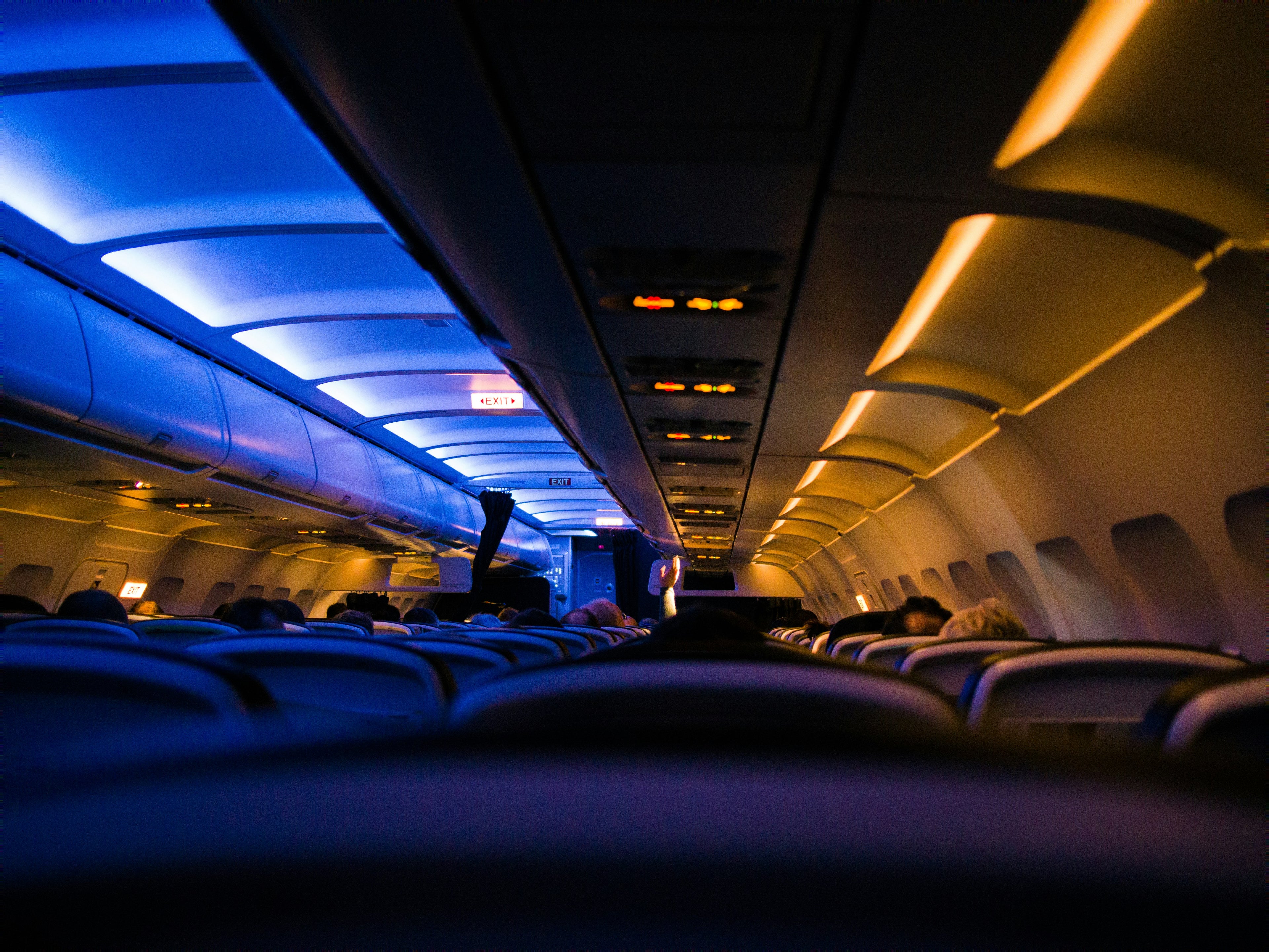 A view from inside an airplane cabin looking down the aisle, with rows of seats, overhead compartments, and blue and warm yellow lighting.