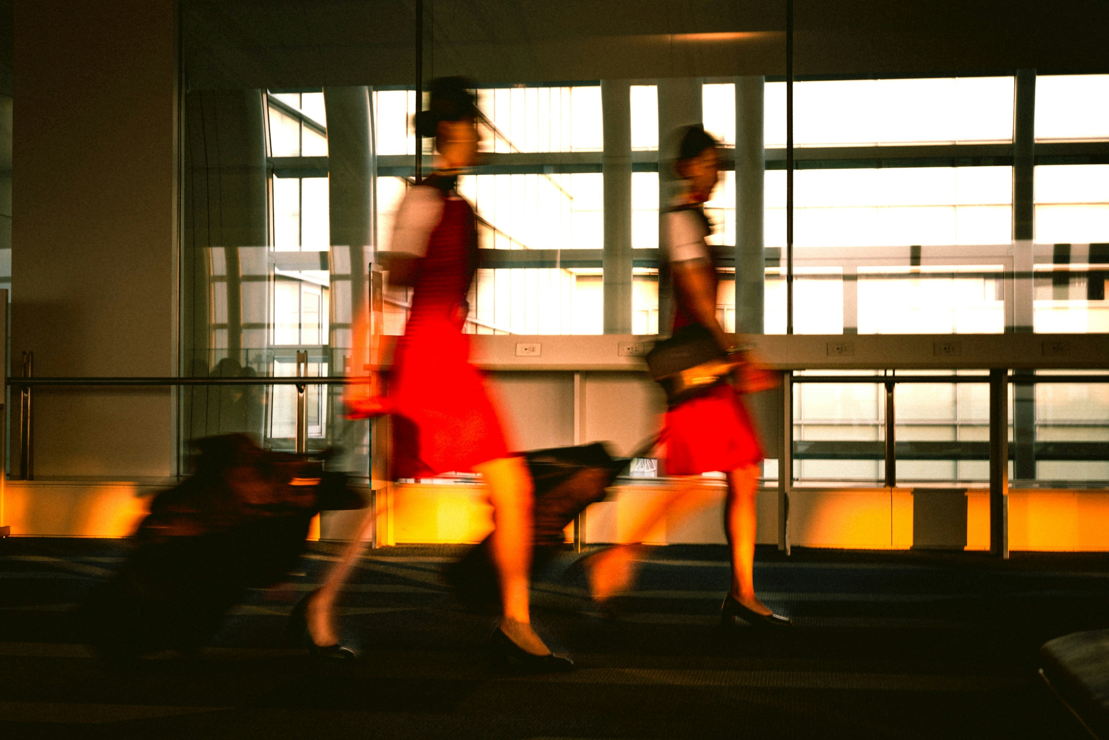 Two women in matching red and white uniforms walking in a motion-blurry indoor setting with large windows and warm orange lighting.